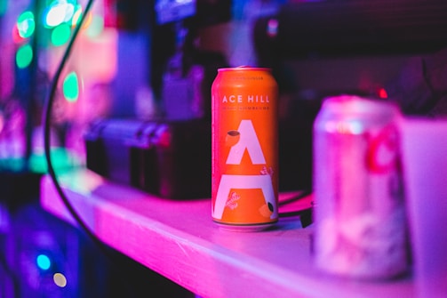 A vibrant scene featuring an orange can of Ace Hill Kombucha prominently placed on a shelf. The background includes various unfocused objects with colorful, blurred lights creating a lively atmosphere. Another can, partially blurred, is visible to the right.