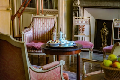An elegant room displays vintage furniture with intricate designs and upholstery featuring pink and white patterns. A round wooden table in the center holds a decorative teapot, cups, and a plate with fruit. A classic fireplace with ornate detailing is visible in the background, complemented by plush chairs arranged around the table.