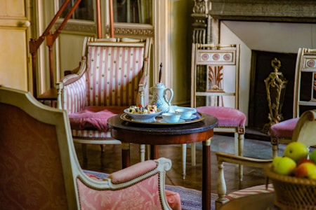 An elegant room displays vintage furniture with intricate designs and upholstery featuring pink and white patterns. A round wooden table in the center holds a decorative teapot, cups, and a plate with fruit. A classic fireplace with ornate detailing is visible in the background, complemented by plush chairs arranged around the table.