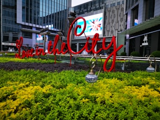A beautifully landscaped urban area with vibrant greenery and a bright red sign reading 'Love in the City'. The background features modern glass buildings and a digital screen displaying colorful images.