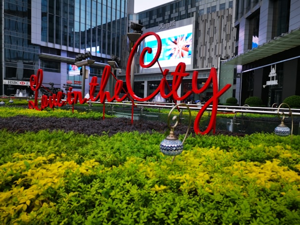 A beautifully landscaped urban area with vibrant greenery and a bright red sign reading 'Love in the City'. The background features modern glass buildings and a digital screen displaying colorful images.