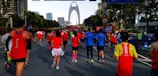 A group of runners participate in a marathon on a city street. They wear colorful athletic clothing, including red and blue shirts. A large modern arch structure is visible in the background. The scene is lively, with spectators and event signs along the route.