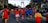 A group of runners participate in a marathon on a city street. They wear colorful athletic clothing, including red and blue shirts. A large modern arch structure is visible in the background. The scene is lively, with spectators and event signs along the route.