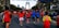 A group of runners participate in a marathon on a city street. They wear colorful athletic clothing, including red and blue shirts. A large modern arch structure is visible in the background. The scene is lively, with spectators and event signs along the route.
