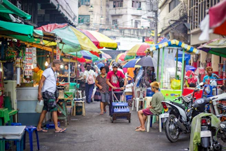 A bustling Laotian market street with vendors and shoppers under colorful awnings.