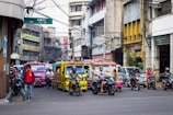 A busy urban street scene with various vehicles, including colorful jeepneys and motorcycles, moving through a congested area. The street is flanked by commercial buildings with signs in multiple languages, and a mix of pedestrians and riders can be seen. Overhead, numerous tangled electrical wires are visible.
