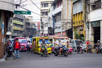 A busy urban street scene with various vehicles, including colorful jeepneys and motorcycles, moving through a congested area. The street is flanked by commercial buildings with signs in multiple languages, and a mix of pedestrians and riders can be seen. Overhead, numerous tangled electrical wires are visible.