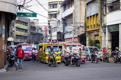 A busy urban street scene with various vehicles, including colorful jeepneys and motorcycles, moving through a congested area. The street is flanked by commercial buildings with signs in multiple languages, and a mix of pedestrians and riders can be seen. Overhead, numerous tangled electrical wires are visible.