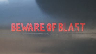 Red text reading 'BEWARE OF BLAST' on a dark metallic surface, suggesting a warning sign typically found on industrial equipment or vehicles.