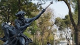 A statue of a mythological figure with a beard holding a trident is set against a backdrop of lush green trees. The setting appears to be an outdoor park area with people walking in the background.