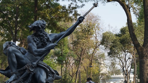 A statue of a mythological figure with a beard holding a trident is set against a backdrop of lush green trees. The setting appears to be an outdoor park area with people walking in the background.
