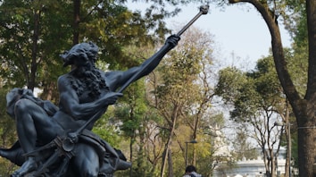 A statue of a mythological figure with a beard holding a trident is set against a backdrop of lush green trees. The setting appears to be an outdoor park area with people walking in the background.