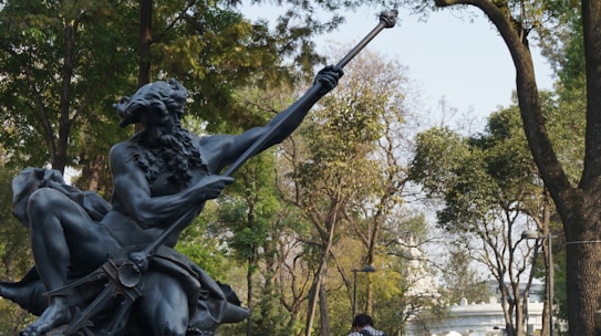 A statue of a mythological figure with a beard holding a trident is set against a backdrop of lush green trees. The setting appears to be an outdoor park area with people walking in the background.