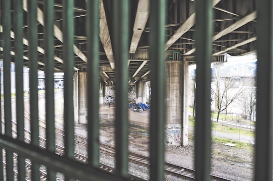 A view through metal bars of a scene underneath an overpass with concrete pillars. There are some tents and belongings suggesting a homeless encampment. Graffiti is visible on the pillars and the ground is gravel with train tracks running through it. Trees and a distant building can be seen in the background.