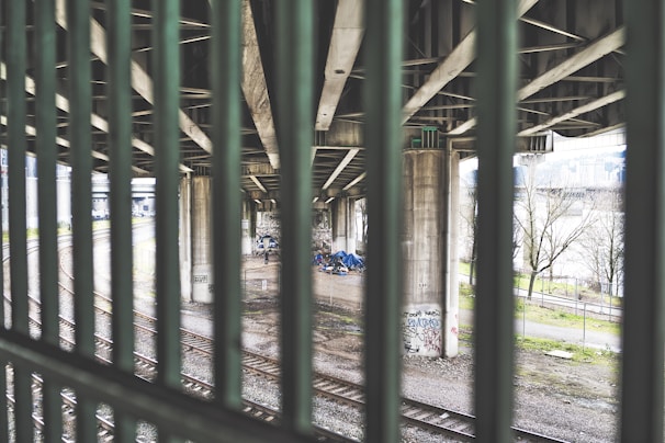 A view through metal bars of a scene underneath an overpass with concrete pillars. There are some tents and belongings suggesting a homeless encampment. Graffiti is visible on the pillars and the ground is gravel with train tracks running through it. Trees and a distant building can be seen in the background.