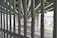 A view through metal bars of a scene underneath an overpass with concrete pillars. There are some tents and belongings suggesting a homeless encampment. Graffiti is visible on the pillars and the ground is gravel with train tracks running through it. Trees and a distant building can be seen in the background.