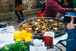 A large pan filled with a seafood dish, likely paella, garnished with lemon wedges, green herbs, and red peppers. In the foreground, there are fresh lemons and greenery, possibly parsley, on the side. There is a carton, possibly of broth or sauce, next to the pan. People in casual clothing are interacting around the table, and a brick wall is visible in the background.