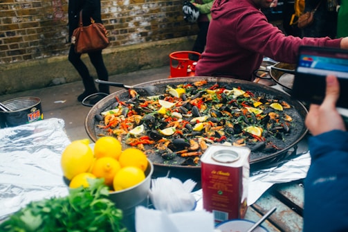 A large pan filled with a seafood dish, likely paella, garnished with lemon wedges, green herbs, and red peppers. In the foreground, there are fresh lemons and greenery, possibly parsley, on the side. There is a carton, possibly of broth or sauce, next to the pan. People in casual clothing are interacting around the table, and a brick wall is visible in the background.