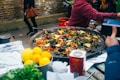 A large pan filled with a seafood dish, likely paella, garnished with lemon wedges, green herbs, and red peppers. In the foreground, there are fresh lemons and greenery, possibly parsley, on the side. There is a carton, possibly of broth or sauce, next to the pan. People in casual clothing are interacting around the table, and a brick wall is visible in the background.