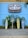 A vibrant surf shop sign with 'Ron Jon Surf Shop' and 'One of a Kind' is displayed against a bright blue wall. Below, three colorful surfboards are arranged vertically with tropical plants in the background. A directional sign points to various beach destinations.