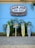 A vibrant surf shop sign with 'Ron Jon Surf Shop' and 'One of a Kind' is displayed against a bright blue wall. Below, three colorful surfboards are arranged vertically with tropical plants in the background. A directional sign points to various beach destinations.