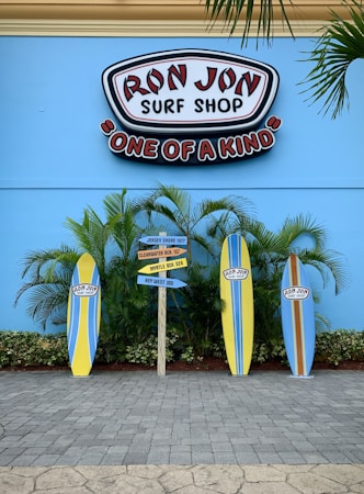A vibrant surf shop sign with 'Ron Jon Surf Shop' and 'One of a Kind' is displayed against a bright blue wall. Below, three colorful surfboards are arranged vertically with tropical plants in the background. A directional sign points to various beach destinations.