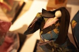 A close-up of a detailed origami figure wearing a colorful, intricate kimono with floral patterns. The figure appears to have long, black hair styled with a bun and a white spherical hairpiece. The focus is primarily on the textures and colors of the origami paper.