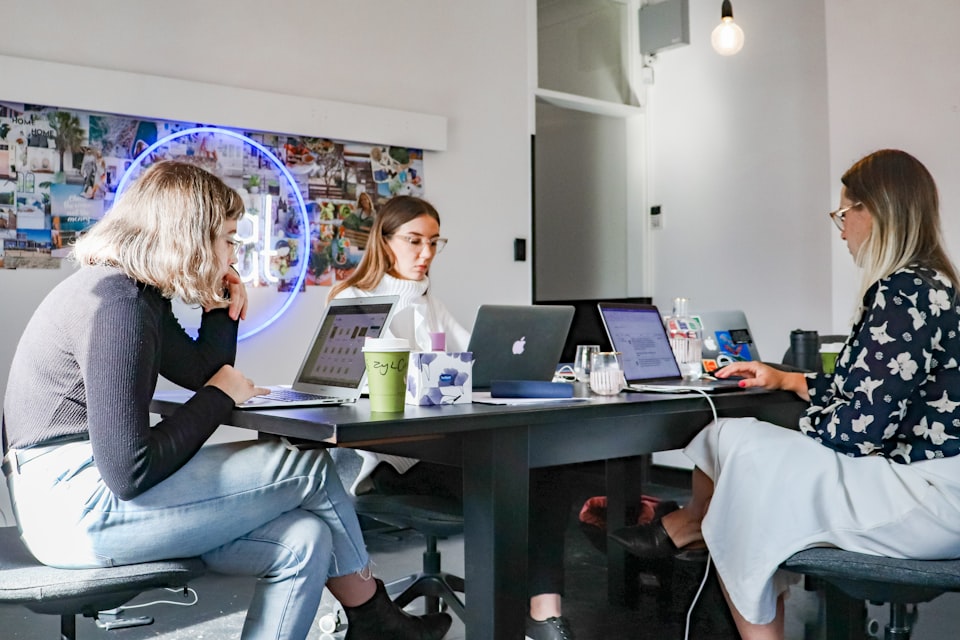 Three women sit at a black table with laptops, beverages, and office supplies.