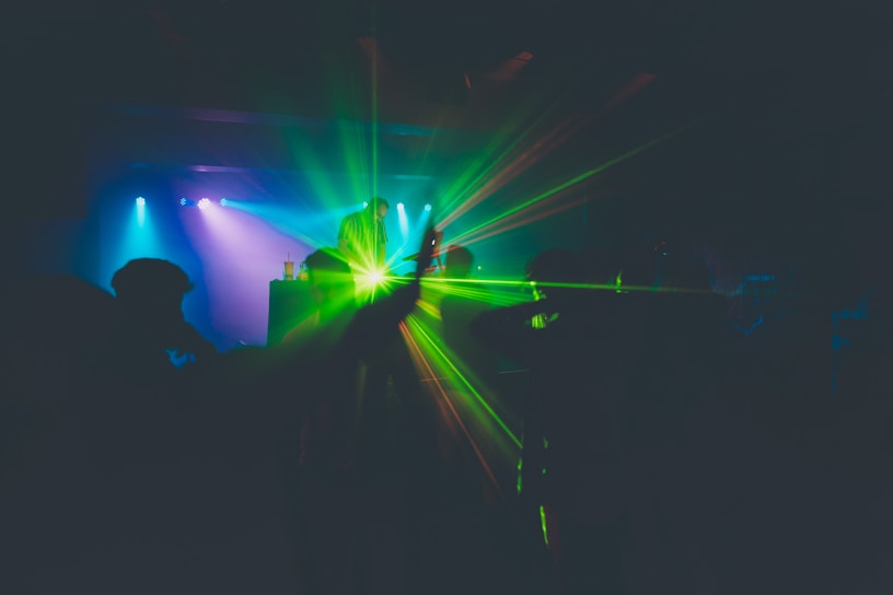 A dimly lit nightclub scene with a DJ performing on stage. Colorful lights and lasers illuminate the space, with green and blue tones creating a vibrant atmosphere. The silhouettes of people dancing are visible, enhancing the energetic mood.