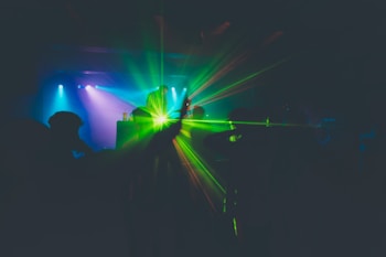 A dimly lit nightclub scene with a DJ performing on stage. Colorful lights and lasers illuminate the space, with green and blue tones creating a vibrant atmosphere. The silhouettes of people dancing are visible, enhancing the energetic mood.