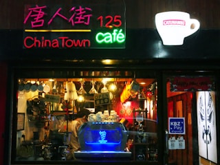 A cozy café with a vibrant neon sign, including the words 'ChinaTown café' and '125'. The decor features hanging utensils and mugs, creating a warm and inviting atmosphere. Inside, a blue-lit coffee machine and various coffee-making equipment are visible. Bright lighting and red accents enhance the vibrant and lively feel of the place.