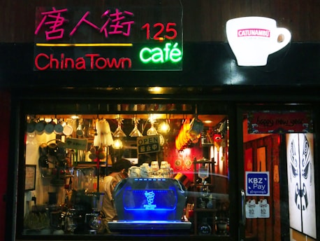 A cozy café with a vibrant neon sign, including the words 'ChinaTown café' and '125'. The decor features hanging utensils and mugs, creating a warm and inviting atmosphere. Inside, a blue-lit coffee machine and various coffee-making equipment are visible. Bright lighting and red accents enhance the vibrant and lively feel of the place.