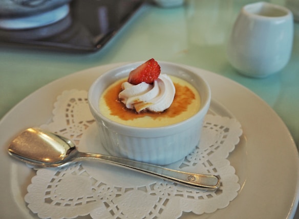 A small white ramekin containing a caramelized dessert topped with a dollop of whipped cream and a slice of strawberry. It is placed on a white plate over a lace paper doily. A silver spoon rests beside the ramekin, and a small white creamer is in the background.
