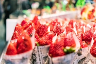 Bright red strawberries are neatly arranged in paper cones on a display. The focus is on the ripe strawberries, showcasing their glossy texture and vibrant color. The background is softly blurred, emphasizing the strawberries in the foreground.