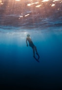 A lone diver glides through the serene underwater environment. Sunlight filters through the water, creating a tranquil ambiance with beams of light illuminating the diver. The scene evokes a sense of peaceful exploration and solitude.