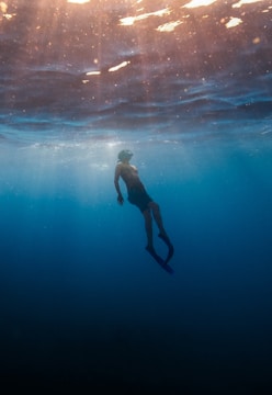 A lone diver glides through the serene underwater environment. Sunlight filters through the water, creating a tranquil ambiance with beams of light illuminating the diver. The scene evokes a sense of peaceful exploration and solitude.