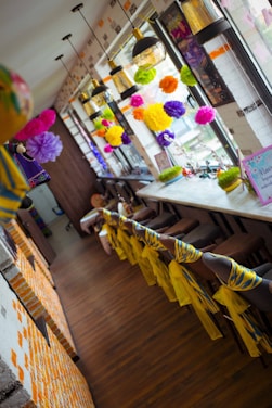 A brightly decorated interior space featuring colorful paper pom-poms hanging near large windows. The long wooden table is lined with stools, each adorned with vibrant yellow and blue fabric. Several pendant lamps with glass shades hang from the ceiling, providing light to the room.