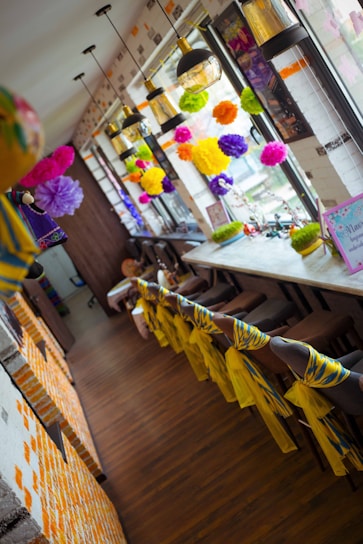 A brightly decorated interior space featuring colorful paper pom-poms hanging near large windows. The long wooden table is lined with stools, each adorned with vibrant yellow and blue fabric. Several pendant lamps with glass shades hang from the ceiling, providing light to the room.