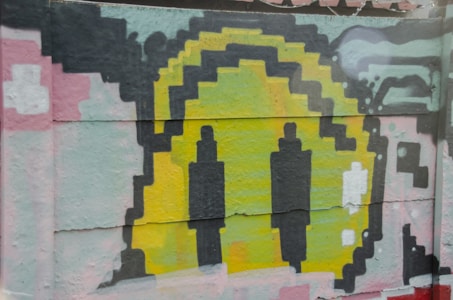 A colorful piece of graffiti art featuring abstract shapes resembling digital or pixelated figures in vibrant hues. The central part is dominated by yellow, surrounded by thick black outlines, and is set against a mix of pastel colors including pink, green, and grey. The art appears on a textured surface, possibly a wall.