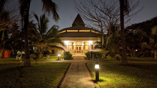 A house is illuminated in the evening, surrounded by palm trees and greenery. A pathway leads up to the front entrance, flanked by lit garden lamps. The sky is dusky, adding a serene atmosphere to the scene.