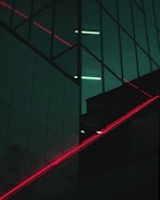 A modern building facade featuring large glass panels with reflections and a grid-like design. Diagonal red neon lights intersect with the dark reflective surfaces, creating contrast against the deep greenish-blue tones.