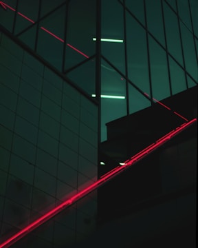 A modern building facade featuring large glass panels with reflections and a grid-like design. Diagonal red neon lights intersect with the dark reflective surfaces, creating contrast against the deep greenish-blue tones.