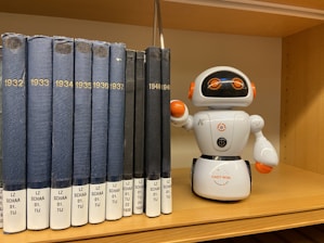 A shelf holding a lineup of blue and black books labeled with years ranging from 1932 to 1941 and a small white robot toy placed next to them. The robot has orange accents and big round eyes with an adorable expression.