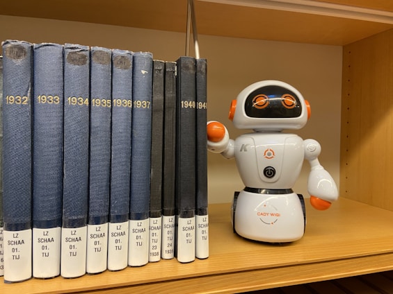 A shelf holding a lineup of blue and black books labeled with years ranging from 1932 to 1941 and a small white robot toy placed next to them. The robot has orange accents and big round eyes with an adorable expression.