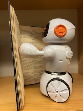 A small, white plastic robot with an orange and black design is positioned next to an open book. It is standing on a wooden surface, possibly a shelf. The robot has wheels for mobility and is facing the book, appearing to be interacting with it.