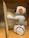 A small, white plastic robot with an orange and black design is positioned next to an open book. It is standing on a wooden surface, possibly a shelf. The robot has wheels for mobility and is facing the book, appearing to be interacting with it.
