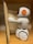 A small, white plastic robot with an orange and black design is positioned next to an open book. It is standing on a wooden surface, possibly a shelf. The robot has wheels for mobility and is facing the book, appearing to be interacting with it.