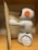 A small, white plastic robot with an orange and black design is positioned next to an open book. It is standing on a wooden surface, possibly a shelf. The robot has wheels for mobility and is facing the book, appearing to be interacting with it.