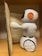 A small, white plastic robot with an orange and black design is positioned next to an open book. It is standing on a wooden surface, possibly a shelf. The robot has wheels for mobility and is facing the book, appearing to be interacting with it.