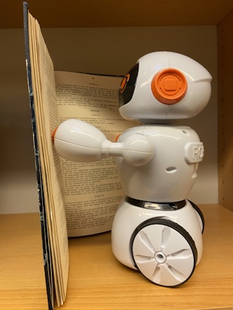 A small, white plastic robot with an orange and black design is positioned next to an open book. It is standing on a wooden surface, possibly a shelf. The robot has wheels for mobility and is facing the book, appearing to be interacting with it.